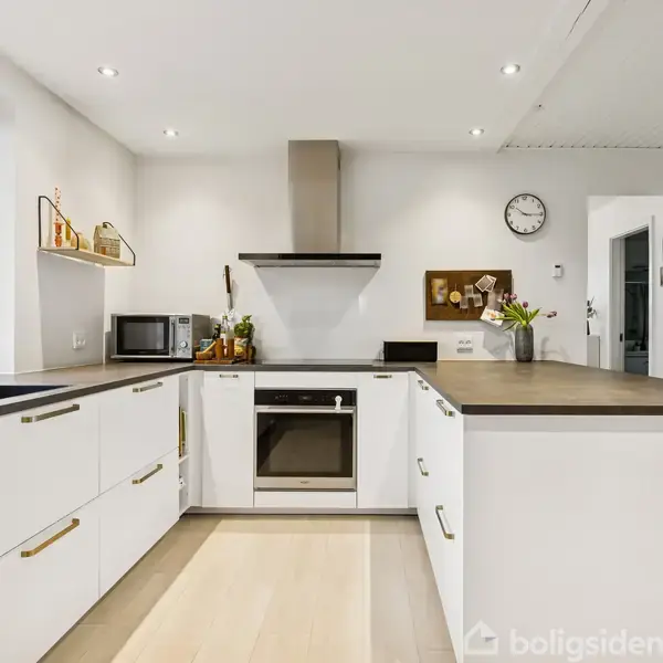 Et moderne køkken med hvide skabe og en mørk bordplade. En indbygget ovn og emhætte er centreret. Et ur hænger på væggen. Vindue til venstre viser lys natur.