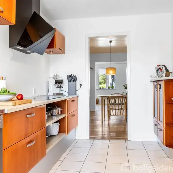 Køkken med træskabe og moderne apparater rummer diverse køkkenredskaber; en åbning fører til spisestue med træstole og et bord, belyst af hængende lampe, og udsigt til en lys fyldt have.