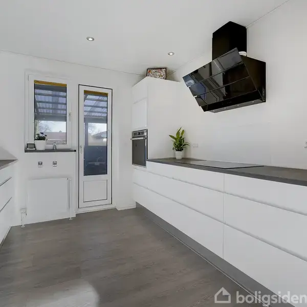 Køkken med hvide skabe strækker sig langs væggene. Emhætte hænger over kogeplade. Dør og vindue fører til udendørs område. Grøn plante og køkkenapparater står på bordpladen.