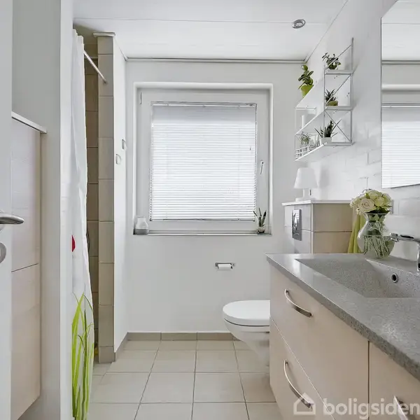 Et moderne badeværelse med en håndvask på en grå bordplade, et spejl på væggen, et toilet ved vinduet og hylder med planter. Flisegulv og lyse vægge.