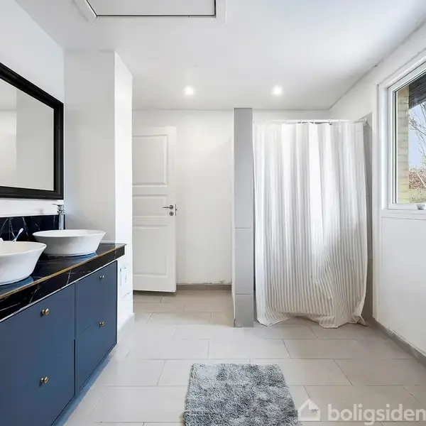 To håndvaske står på en sort marmorbordplade i et lyst badeværelse. Ved siden af er der et bruseområde med et hvidt forhæng. Store spejle og et vindue kompletterer rummet.