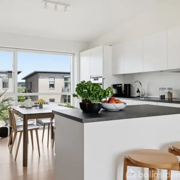 Et lyst køkken med et sort køkkenbord, dekoreret med grøntsager og frugt. Ved siden af står en spisestue med fire stole, omgivet af store vinduer og grønne planter.
