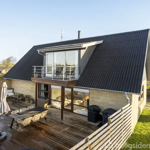 Et moderne hus med stor træterrasse fyldt med udemøbler, der står i solrig have. Store vinduer og altan ses på førstesalen, omgivet af andre huse i nabolaget.