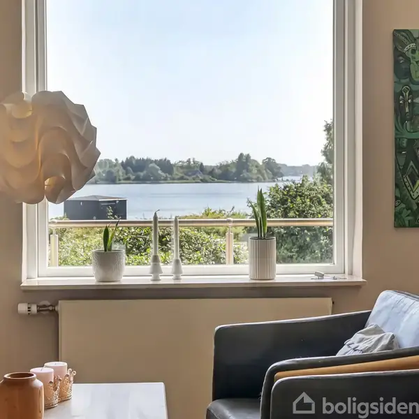 En stue med lænestol og sofabord, hvorfra der er udsigt til en sø gennem et stort vindue. Bordet har pynteting, og der hænger billeder på væggene.