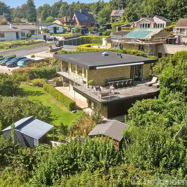 To-etagers hus med stor terrasse er omgivet af en frodig have med træer og buske. I baggrunden ses en række andre huse og en parkeringsplads med biler.