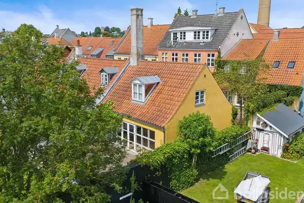 Et gult hus med rødt tag ses fra oven, omgivet af en grøn have og andre huse. I baggrunden er der en blå himmel med få skyer.