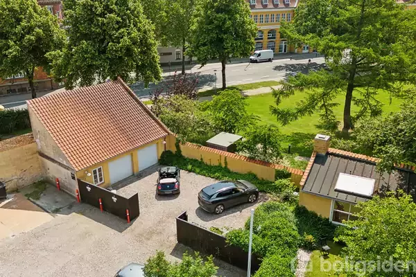 To biler holder parkeret foran en gul bygning med rødt tegltag. I baggrunden ses en offentlig græsplæne med træer, og en vej med biler kørende forbi.