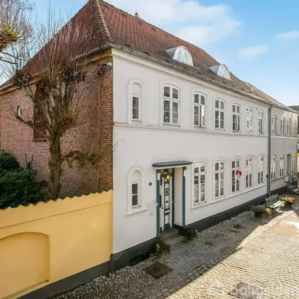Et charmerende hvidt murstenshus med buede vinduer står langs en brostensbelagt gade. En lille gul mur omslutter en have på siden. Klart sollys skaber en hyggelig stemning.