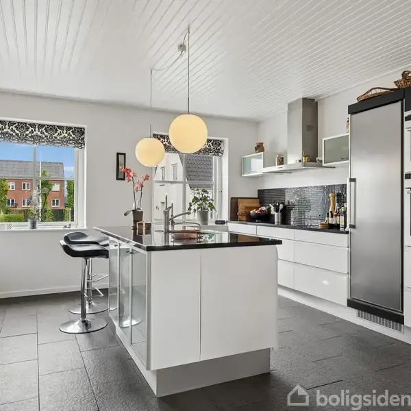 Køkken med hvidt interiør og sort gulv. Bord i midten omgivet af barstole. Vinduer med udsigt til bygning udenfor. Moderne apparater indbygget i væggen.