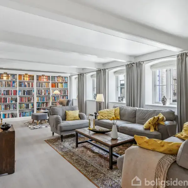 Stue med grå sofaer og gul puder i aktion som afslapningsområde. Reol fyldt med bøger skaber intellektuelt miljø. Store vinduer med lyse gardiner tilføjer lys og rummelighed.