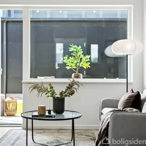 Stue med grå sofa ved siden af et rundt sofabord med planter og et stearinlys. Store vinduer bagved giver udsigt til en åben dør og en udendørs terrasse.