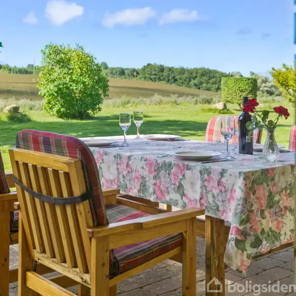To træstole med farverige hynder, står ved et bord med blomstrede duge og vinglas. Bordet er pyntet med røde blomster, og det hele befinder sig på en terrasse med udsigt over marker og træer.
