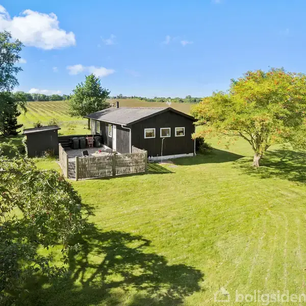 En sort træhytte med en lille gårdhave står på en stor græsplæne omgivet af træer. Skyfri blå himmel og marker ses i baggrunden.