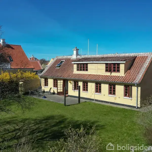 Et gult hus med rødt tegltag står stille i en have omkranset af buske og træer, med klart blå himmel i baggrunden.