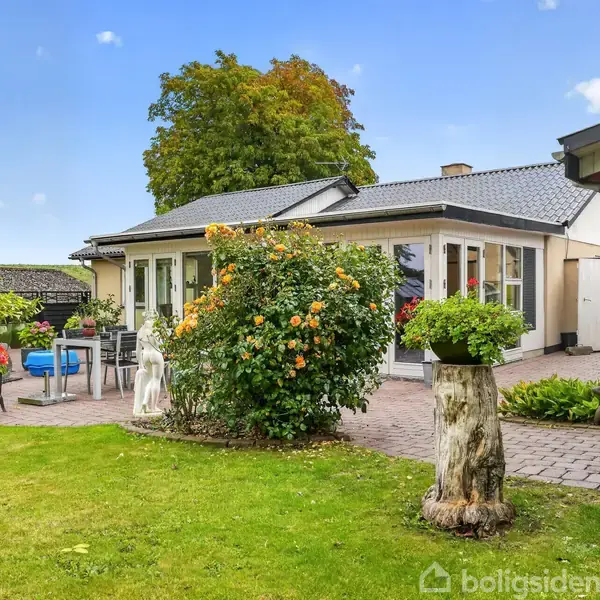 En lyserød murstensvilla med hvidrammede vinduer står i en velplejet have med blomster og grøn græsplæne. Et flisebelagt område har havemøbler og en grill.