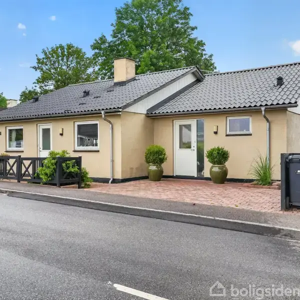 Et enfamilieshus med beige facade og sort tag står langs en vej. Foran er der fliser og to grønne planter i krukker. I baggrunden er der træer og blå himmel.