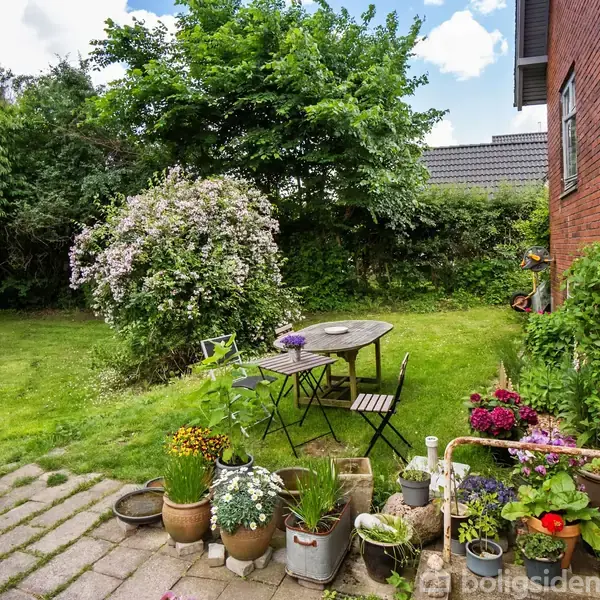 Et rundt metalbord med to stole står på en terrasse. Omkring dem er blomsterkrukker i en have med græsplæne og buske nær en murstensbygning.