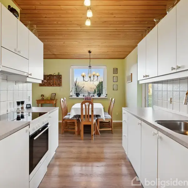 Et køkken med hvide skabe udstyret med ovn og vask, fører til en spiseplads med træmøbler under en lysekrone. Trægulv og lofter skaber en hyggelig atmosfære.