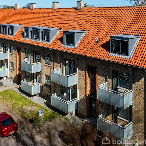 En række murstensbygninger med flere altaner. Biler holder parkeret på vejen ved siden af bygningen. Taget er dækket af røde tegl. Buske og træer omgiver området.
