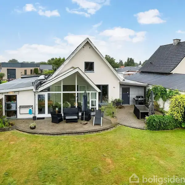 Et hvidt hus med skrå tag og store vinduer vender ud mod en grøn have med terrasse. Havemøbler står på terrassen, omkranset af veltrimmet hæk og buske.