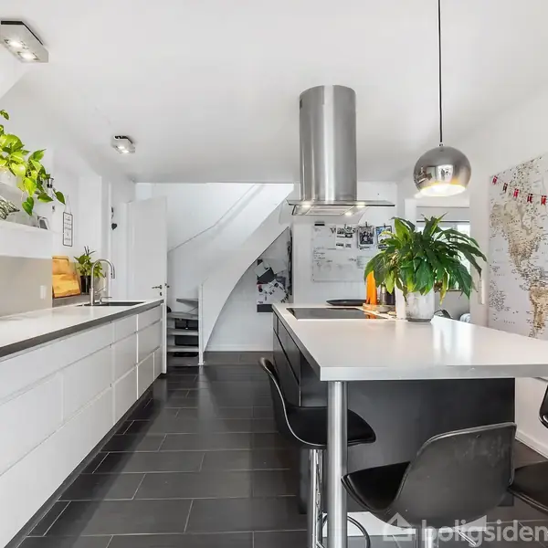 Køkken med moderne design; et langt bord har barstole og planter. Hvide skabe langs væggen, ovn indbygget, verdens kort på væggen. Lysindfald fra loftlamper.