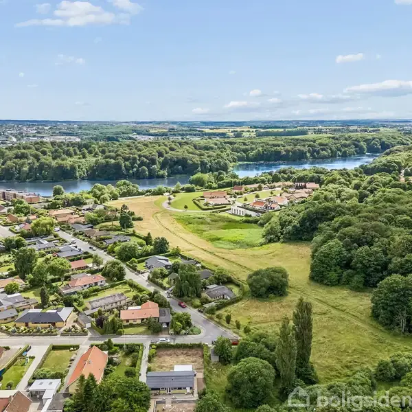 Luftfoto viser et boligkvarter med parcelhuse, markeret med en blå pil, omgivet af frodige skove og en stor sø i baggrunden.
