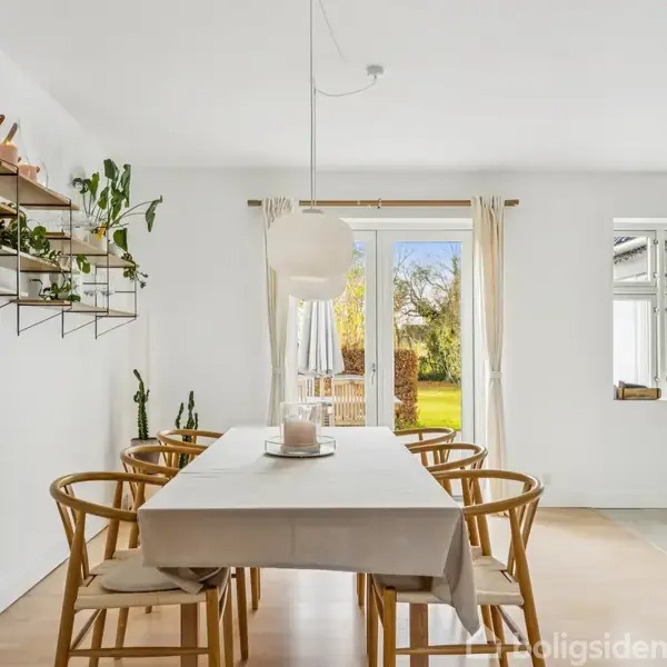 Et spisebord med seks stole står midt i et lyst rum. Bordet er dækket med en hvid dug, og ovenover hænger en lampe. Væggen har hylder med planter og billeder. En åben dør viser en have udenfor.