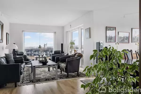 Stue med sorte lædersofaer og -stole omkring et sofabord; vinduer giver udsigt til byen. Grønne planter dekorerer rummet, der er lyst og rummeligt med parketgulv.