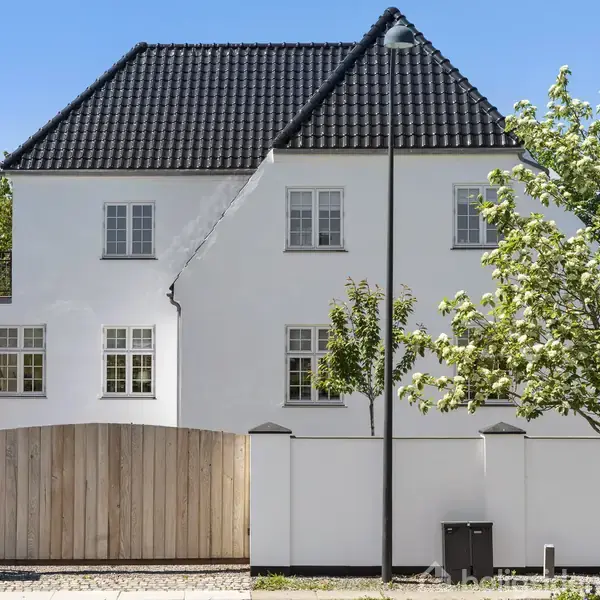 Hus med hvid facade og sort tegltag står roligt bag en hvid mur med træport. Et gadelys og et træ præger forgrunden, omgivet af grønne buske og blå himmel.