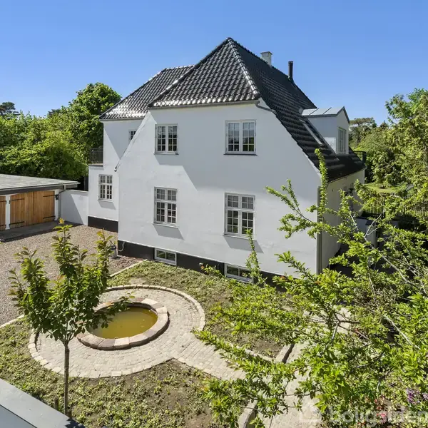Hvidt hus med sort tag står stille. I haven ses en rund dam omgivet af flisebelægning. Omgivelserne består af grønne træer og buske.