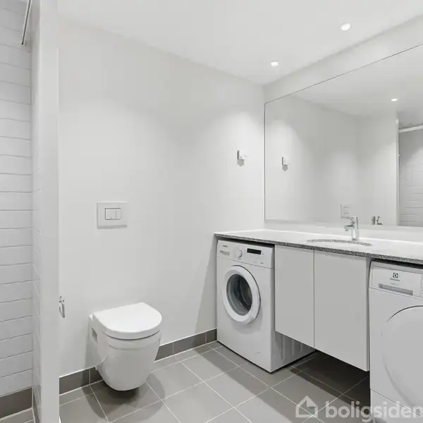 Toilet hænger på væggen over grå fliser, ved siden af en brusekabine. En vaskemaskine og opvaskemaskine er indbygget under en spejlbeklædt væg i et moderne badeværelse.