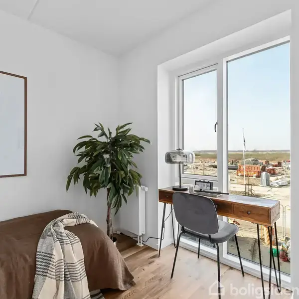 En stol står ved et skrivebord med en lampe og computer foran et vindue. Ved siden af er en grøn plante, og der er udsigt til en byggeplads udenfor.