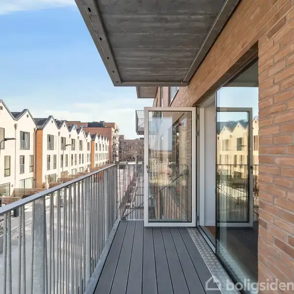 Balkon med åbent vindue, der fører fra en murstensbygning. Udsigt over en gade med moderne rækkehuse og en klar blå himmel.