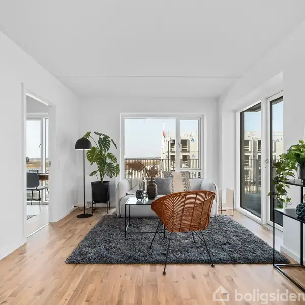 Stuen indeholder en grå sofa på et mørkt tæppe, omgivet af en stueplante og et glasvindue mod en balkon. Et sidebord med lampe og dekorative genstande står ved siden af.