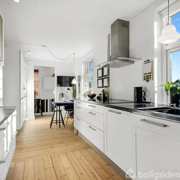 Et moderne køkken med hvide skabe strækker sig langs væggene. En sort bordplade understreger indretningen. I baggrunden ses et spiseområde med stole ved et bord, omgivet af store vinduer.