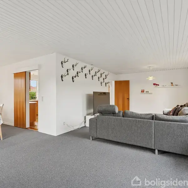 Grå sofa står i en stue med hvidt loft og gulvtæppe, ved siden af et fjernsyn. Et spisebord med stole ses til venstre. Rummet er lyst af store vinduer.