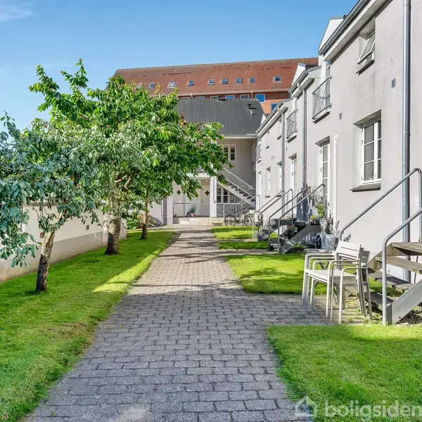 Gangsti forbinder rækkehuse af lysegrå facader med døre og trapper; træer pryder græsområdet ved siden af, stol og bord ved en trappe. Klar blå himmel ses i baggrunden.