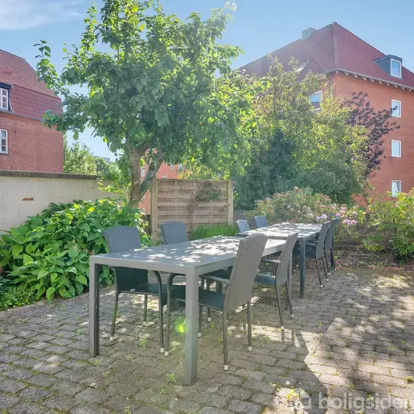 En lang havebord er omgivet af sorte stole i en gårdhave med flisebelægning. Et træ og grønne buske skaber en hyggelig atmosfære foran røde murstensbygninger.