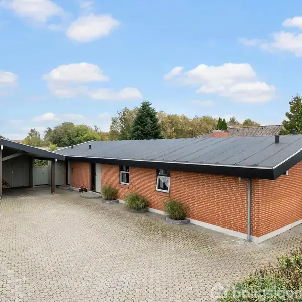 Et rødt murstenshus står stille på en flisebelagt gårdsplads. Til venstre er der en carport med en bil. Omgivet af grønne buske og træer under en blå himmel.