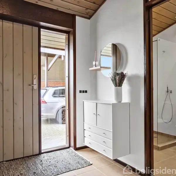 En entre med en hoveddør til venstre, hvor en bil er synlig udenfor. Et hvidt opbevaringsmøbel står mod væggen med en vase med græs. Til højre er et badeværelse med toilet og bruser. En rund spejl hænger over møblet. Ingen tekst fundet.