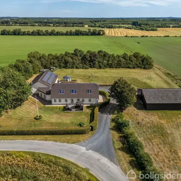 En stor landejendom ligger omgivet af grønne marker og træer. Huset har en grusvej, der fører til hovedindgangen. Ved siden af er en stor lade.