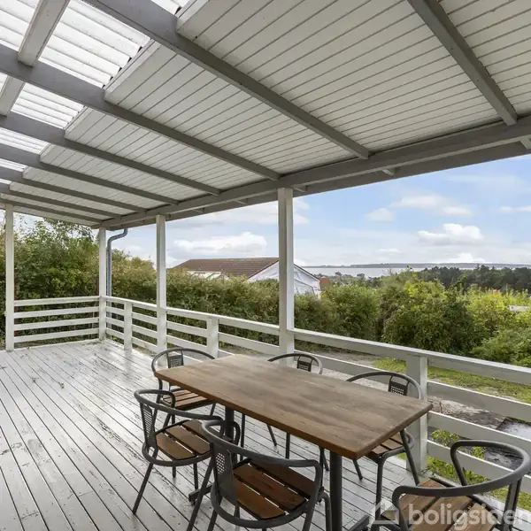En træterrasse med tag og hvidmalet rækværk har et bord med seks stole. I baggrunden ses en have med grønne buske og udsigt til bakker under en klar himmel.