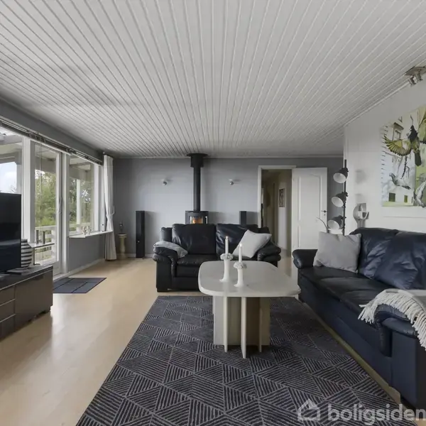 En stue med sorte lædersofaer og et sofabord står i et rum med trægulv, stor fladskærms-TV og en brændeovn i hjørnet. Billedkunst hænger på væggen.