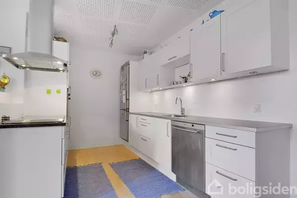 Hvidt køkken med opvaskemaskine og indbyggede skabe, belyst af loftslamper. Køleskab i baggrunden ved siden af en vægmonteret ur. Blå tæpper på trægulvet.