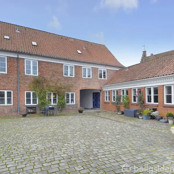 Bygning står stille i gårdsplads med brosten, omkranset af murstensvægge og rødt tegltag. Potteplanter pynter langs gavlen, og himlen er blå med få skyer.