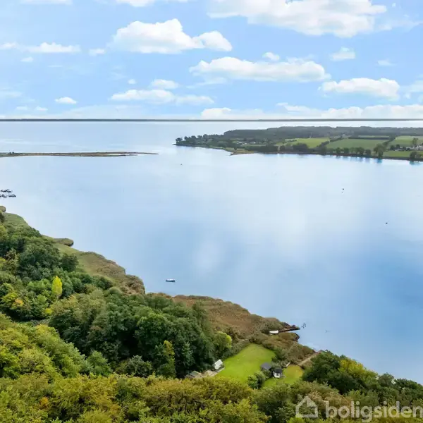 En stor sø breder sig ud i midten, omgivet af frodig skov og en vej til venstre. Landskabet bagved præsenterer åbne marker og skovområder under en skyet himmel.