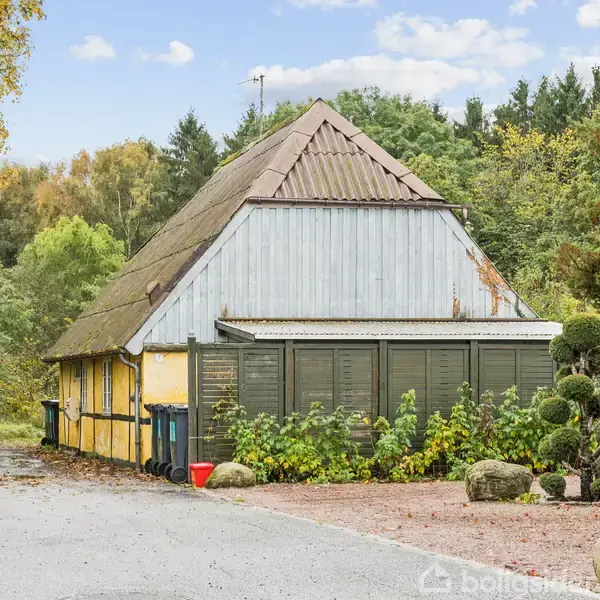 En gul og grå bygning med skråt tag står omgivet af frodig vegetation og træer. En asfalteret vej og buske med omhyggeligt trimmet buksbom sætter scenen.