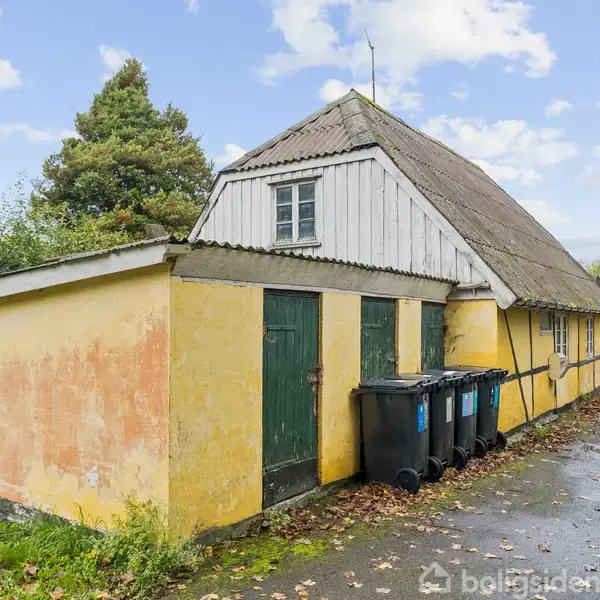 Et gulmalet hus med skråt tag og grønne døre står i en grøn, åben have. Sorte skraldespande er placeret langs husets væg. Træer omgiver området.
