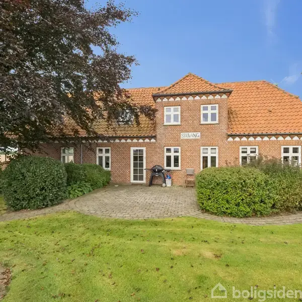Et rødt murstenshus med rødt tegltag og hvide vinduer står på en velplejet plæne med flisebelagt gangsti. Skiltet "Sving" hænger over døren. Fliseareal rundt om huset.