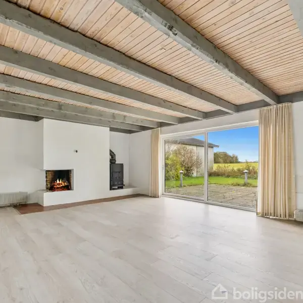 Stue med åbent ildsted brænder roligt. Store vinduer åbner ud mod en grøn have. Lys trægulv og synlige loftsbjælker giver et rummeligt indtryk. Flyttekasser står i hjørnet.